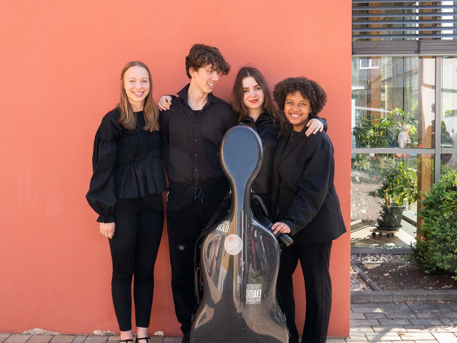 Vier Jugendliche vor einer orangenen Wand, in der Mitt ein Cellokasten.